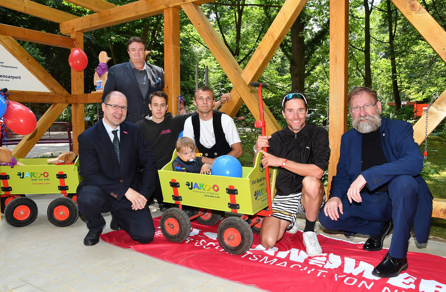 Triathlet ausnahmsweise auf vier Rädern - der Rostocker Ironman Andreas Raelert (2.v.re.) kam zufällig während der Einweihung des Carports mit seinem Sohn Theo (2) im Bollerwagen vorbei - hier mit Vizepräsident Jens Meinert (v. li.), Bau-Präsident Rainer Bauer, die Zimmererlehrlinge Wiliam Tomaszewski und Paul Blohm und Zoodirektor Udo Nagel (re.). 